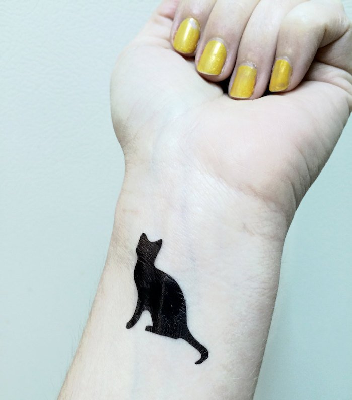 कलाई पर एक बिल्ली टैटू के लिए बहुत अच्छा विचार - यहाँ एक हाथ, पीले नेल पॉलिश और एक छोटी सी काली बिल्ली है